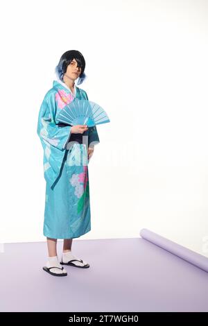 giovane uomo in kimono blu e parrucca in posa con ventola a mano su tappeto viola e sfondo bianco, cosplay Foto Stock