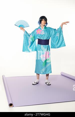 uomo espressivo in kimono blu e parrucca in posa con ventola a mano su tappeto viola e sfondo bianco Foto Stock