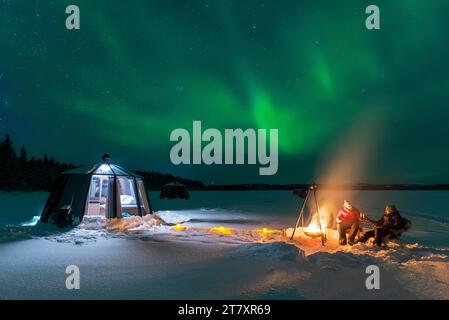Vista notturna di due persone e cena all'aperto intorno a un falò vicino all'igloo di vetro illuminato con l'aurora boreale Foto Stock