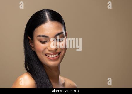 Ritratto di donna birazziale con capelli scuri, sorridente e trucco naturale su sfondo marrone Foto Stock