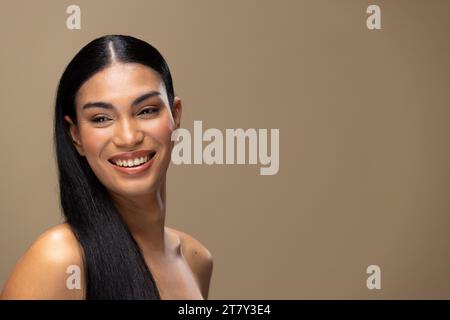 Ritratto di donna birazziale con capelli scuri, sorridente e trucco naturale su sfondo marrone Foto Stock