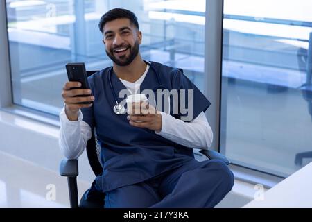 Ritratto del medico maschio birazziale felice utilizzando lo smartphone e tenendo tazza di caffè in ufficio ospedale Foto Stock