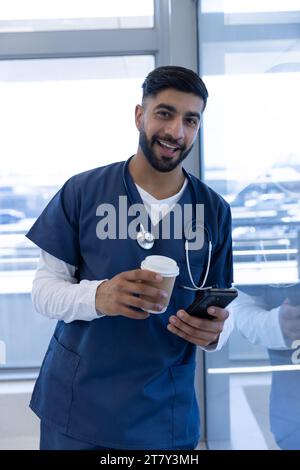 Ritratto del medico maschio birazziale felice utilizzando lo smartphone e tenendo tazza di caffè in ufficio ospedale Foto Stock