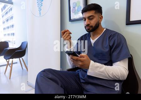 Medico maschio birazziale che utilizza lo smartphone nella sala d'attesa dell'ospedale Foto Stock