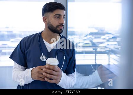 Medico maschio birazziale che utilizza lo smartphone e tenendo la tazza di caffè in ufficio ospedaliero Foto Stock