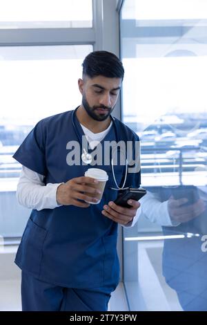 Medico maschio birazziale che utilizza lo smartphone e tenendo la tazza di caffè in ufficio ospedaliero Foto Stock