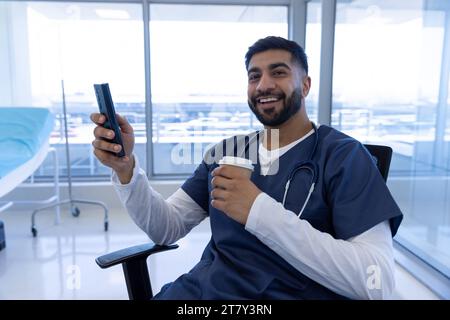 Ritratto del medico maschio birazziale felice utilizzando lo smartphone e tenendo tazza di caffè in ufficio ospedale Foto Stock