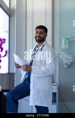 Ritratto di felice medico birazziale maschio che legge documento in soleggiata stanza d'ospedale Foto Stock