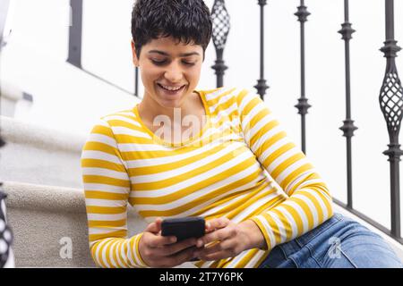 Felice donna birazziale che usa lo smartphone seduto sulle scale a casa Foto Stock