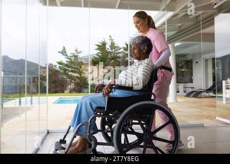 Infermiera caucasica felice e paziente anziana afroamericana in sedia a rotelle Foto Stock