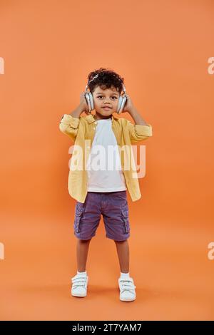 un ragazzo afro-americano allegro e riccio che ascolta musica con cuffie wireless su sfondo arancione Foto Stock