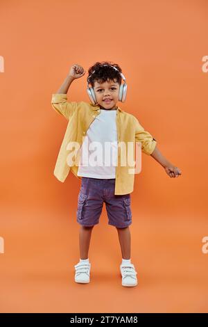 un ragazzo afro-americano sorridente e riccio che ascolta musica con cuffie wireless su sfondo arancione Foto Stock