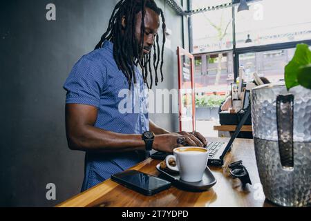 Uomo africano con dreadlocks, barba e camicia, uomo d'affari autonomo, in piedi in una caffetteria che lavora al suo computer portatile e beve caffè, spazio per le copie Foto Stock