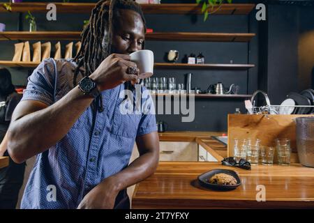 uomo nero con dreadlocks con barba e abiti informali, cliente al ristorante che si piega al bancone bevendo e gustando una tazza di caffè Foto Stock