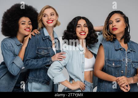 amiche multietniche attraenti e felici in denim indossano in posa grigia, diversità e moda Foto Stock