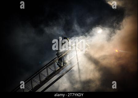 Un pompiere si arrampica su una lunga scala da un camion in azione per combattere uno spettacolare blaise presso il legname RK Miles a Montpelier, VT, New England, USA. Foto Stock