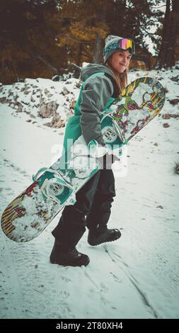 Una giovane donna adulta in attrezzatura invernale che cammina su un terreno innevato, portando uno snowboard tra le braccia Foto Stock