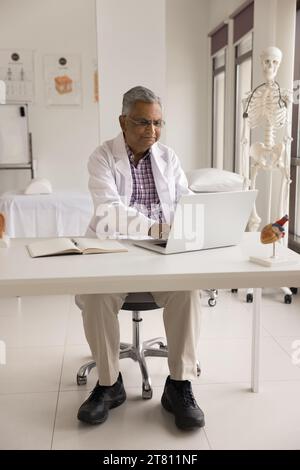 Positivo vecchio indiano medico uomo che lavora al tavolo dell'ufficio dell'ospedale Foto Stock