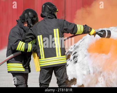 due vigili del fuoco in uniforme con caschi protettivi spengono il fuoco con schiuma bianca Foto Stock