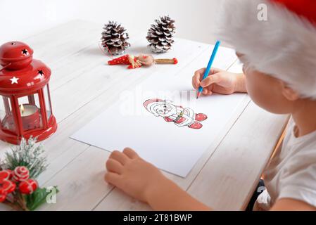 Giovane ragazza che disegna Babbo Natale su carta con un cappello festivo. Decorazioni natalizie su un tavolo di legno bianco. Concetto creativo per le vacanze Foto Stock