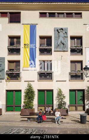 Donne sedute su una panchina al sole nella piazza principale della città di Castellon. Palazzo amministrativo municipale. Bandiera Ucraina. Non alla guerra, Spagna Foto Stock