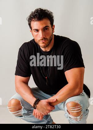 Un uomo accovacciato a terra che indossa jeans strappati, in studio, su sfondo bianco Foto Stock