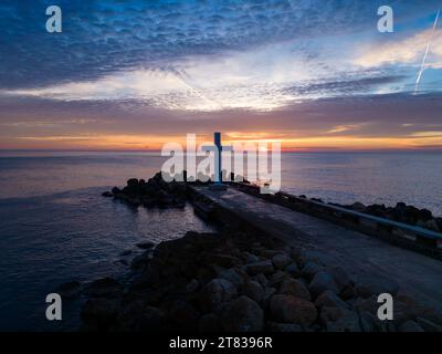 Un volo intorno a una croce cristiana la mattina presto all'alba. La grande croce sorge sul bordo di un frangiflutti sulla costa del mare. Foto Stock