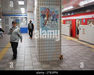 TOKYO, GIAPPONE - 10 novembre 2023: Persona che usa lo smartphone e cammina sulla piattaforma della stazione della metropolitana di Tokyo con un poster che avverte dei pericoli di un atto del genere. Foto Stock