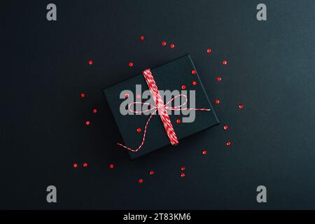 Regalo in scatola nera e coriandoli rossi su sfondo nero. Concetto di Natale. Vista dall'alto, base piatta. Foto Stock