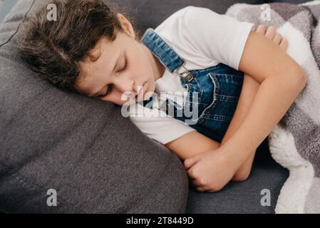 Un piccolo ragazzo caucasico turbato ha la febbre. Concetto di salute, malattia, malattia, raffreddore comune, trattamento. Primo piano della bambina malata che giace da sola su gr Foto Stock