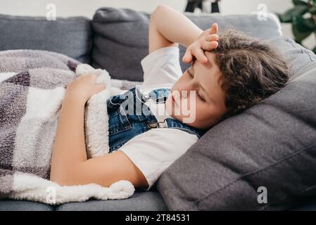 Primo piano di bambino malato malsano sdraiato da solo sul divano sotto coperta in soggiorno a casa. Un piccolo ragazzo caucasico turbato ha la febbre. Concetto di Foto Stock