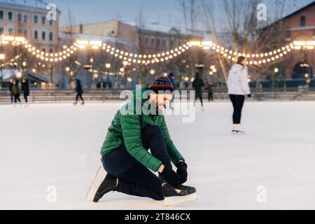 Uomo che lega i pattini di ghiaccio su una pista illuminata durante le feste al crepuscolo, con pattinatori dietro. Foto Stock