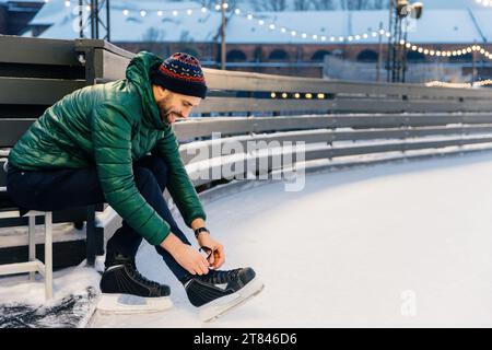 Uomo concentrato che si allaccia sui pattini di ghiaccio in una pista innevata, con luci calde sullo sfondo Foto Stock