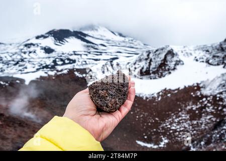 Mano turistica che tiene la pietra lavica sul cratere dell'Etna, Sicilia, Italia. Formazione di roccia lavica vulcanica calda. Paesaggio di crateri pendii ricoperti di neve Foto Stock