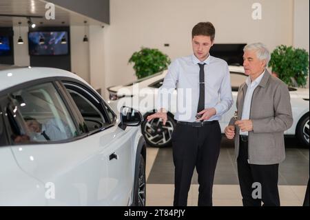 Un venditore mostra un'auto a un uomo anziano in uno showroom. Foto Stock