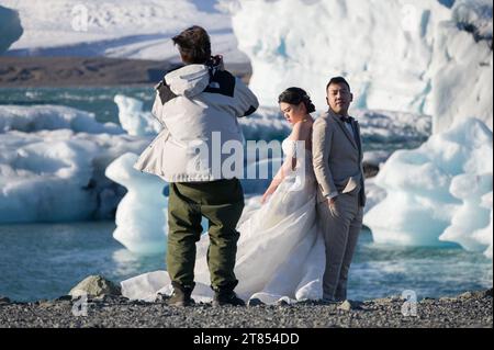 Una coppia asiatica/cinese che indossa un abito da sposa e un abito da sposa posa per foto alla laguna di Jokulsarlon con iceberg sullo sfondo, Islanda Foto Stock