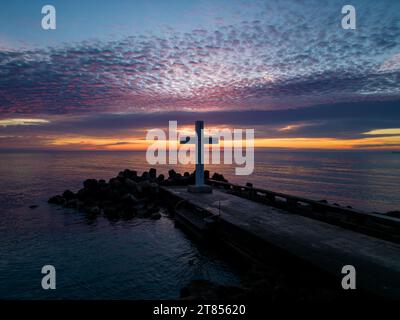 Un volo intorno a una croce cristiana la mattina presto all'alba. La grande croce sorge sul bordo di un frangiflutti sulla costa del mare. Foto Stock