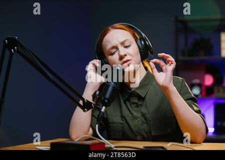 Giovane donna adulta moderna che indossa cuffie per registrare podcast o trasmettere in studio domestico. Foto Stock