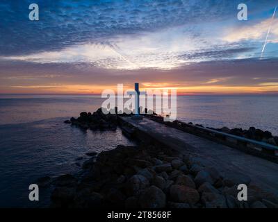 Un volo intorno a una croce cristiana la mattina presto all'alba. La grande croce sorge sul bordo di un frangiflutti sulla costa del mare. Foto Stock