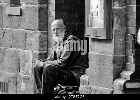 Brecon, Powys, Galles 22 ottobre 2023: Vecchio senzatetto dai capelli grigi con barba piena seduto all'ingresso degli uffici del Consiglio della Guild Hall. Foto Stock