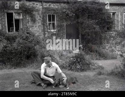 Anni '1950, storico, un gentiluomo con il suo cane da compagnia davanti a un cottage di campagna inglese, che gli dà una sorpresa, un maiale, Inghilterra, Regno Unito. Foto Stock