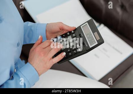 Foto ritagliata di una contabile in camicia blu calcola i costi della calcolatrice, conta i soldi, il budget aziendale, analizza i report finanziari, controlla la fattura Foto Stock