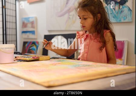 Bella ragazza caucasica di talento, piccola artista con lunghi capelli ricci, vestita in abito rosa, che mescola acquerelli, tiene pennello e disegna pi Foto Stock