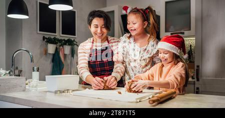 Ridendo bambine in cappello rosso di Santa con madre impastare pasta di pan di zenzero di Natale insieme in cucina domestica. Felice vacanza preparazione e bambino Foto Stock