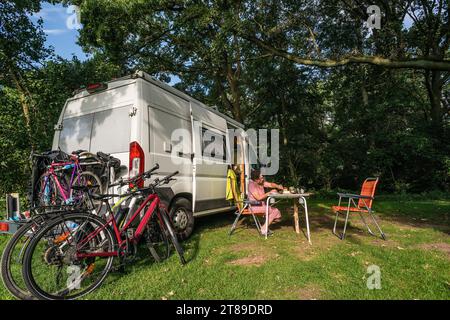 Ein camper mit Fahrrädern auf dem Heckträger steht in idylischer Umgebung im Sommer auf einem Camperstellplatz, eine Frau sitzt am Campingtisch. Spreewald, Brandeburgo, Deutschland. Unterwegs im sommerlichen Spreewald *** Un camper con biciclette sul portapacchi posteriore si trova in un ambiente idilliaco in estate, in un campeggio, una donna siede al tavolo da campeggio Spreewald, Brandeburgo, Germania sulla strada in estate Spreewald Credit: Imago/Alamy Live News Foto Stock