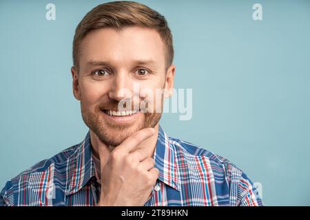 Ritratto uomo barbuto caucasico che guarda la fotocamera sorridente toccando il mento su sfondo blu. Foto Stock