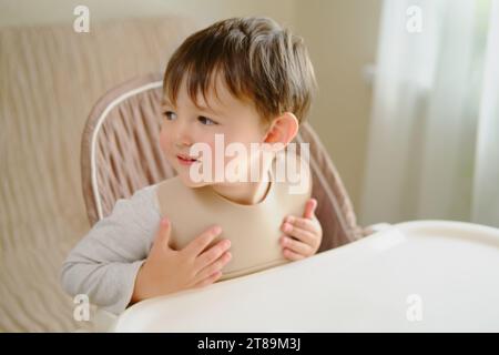 Il bambino attende con impazienza il cibo seduto al tavolo in un bavaglino. Ragazzo di due anni (di due anni) Foto Stock