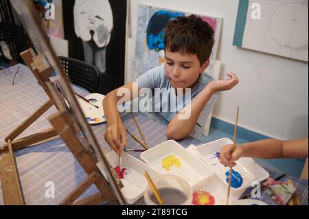 Autentico e sicuro, felice ragazzo adolescente, studente di scuola elementare dipinge quadri su tela, durante le lezioni di arte creativa e artigianato. Persone. Leisu Foto Stock