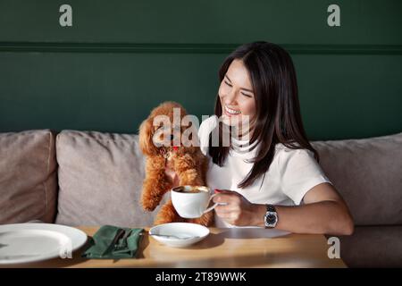 Bella ragazza sorridente con un piccolo cane in braccio seduto al tavolo del ristorante con una tazza di caffè. Donna con capelli scuri in caffetteria con animale domestico su sfondo scuro Foto Stock