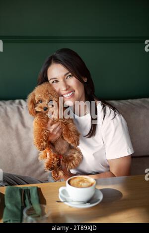 Bella donna sorridente tiene in mano un piccolo cane siede al tavolo del ristorante con una tazza di caffè. Donna dai capelli scuri nella caffetteria con barboncino in miniatura Foto Stock